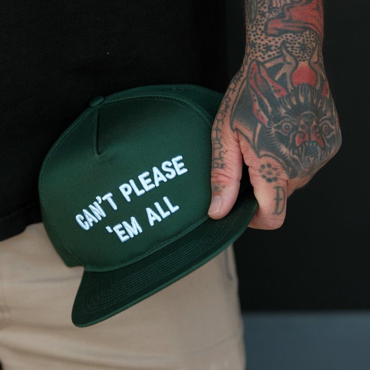 A close-up of a person holding a dark green snapback cap with white embroidered text that reads &