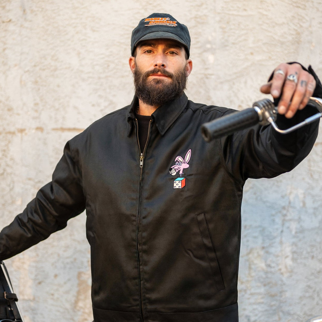 Man wearing a black jacket with a logo and a cap, holding a bicycle.