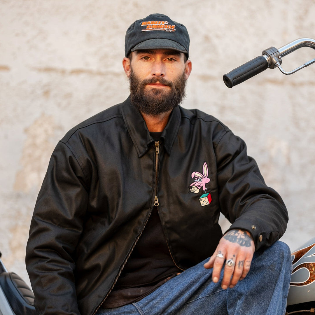 Man wearing a black jacket and cap sitting on a motorcycle.