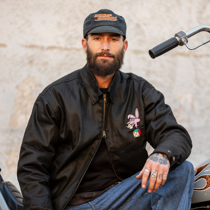 Man wearing a black jacket and cap sitting on a motorcycle.