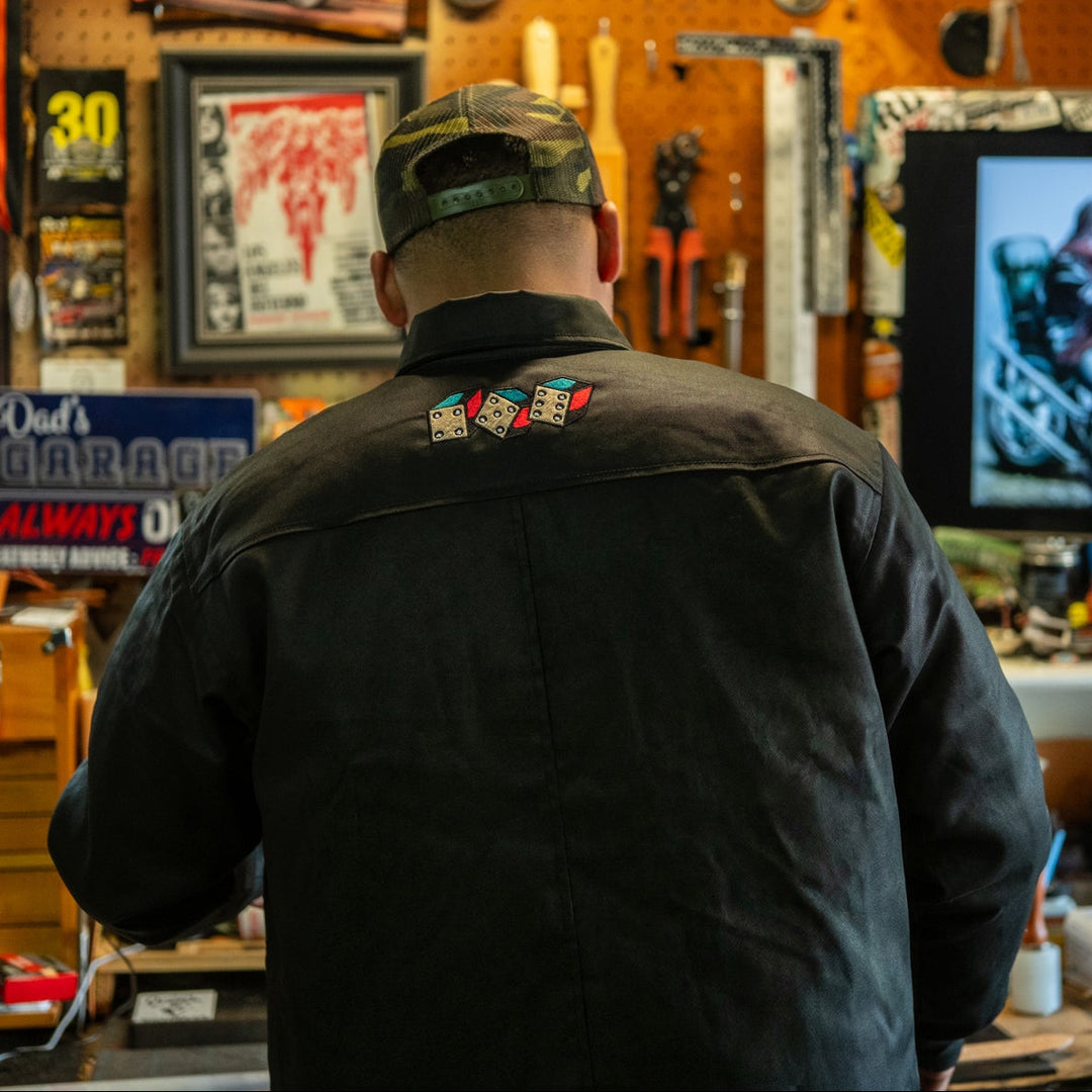 Person wearing a black jacket with patches in a workshop setting, facing a television screen.