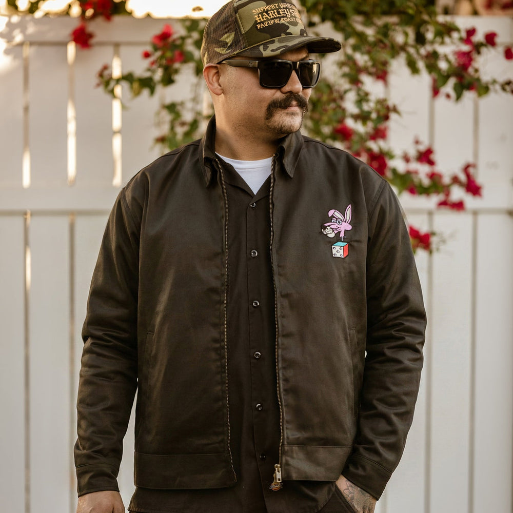 Man wearing a dark jacket with a logo, camouflage cap, and sunglasses against a white fence with red flowers.