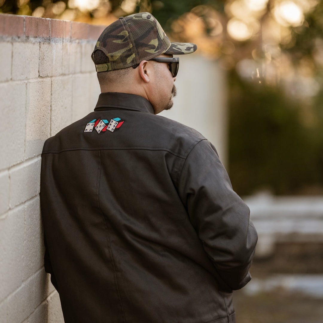 Man wearing a black jacket with patches, camouflage cap, and sunglasses leaning against a wall.