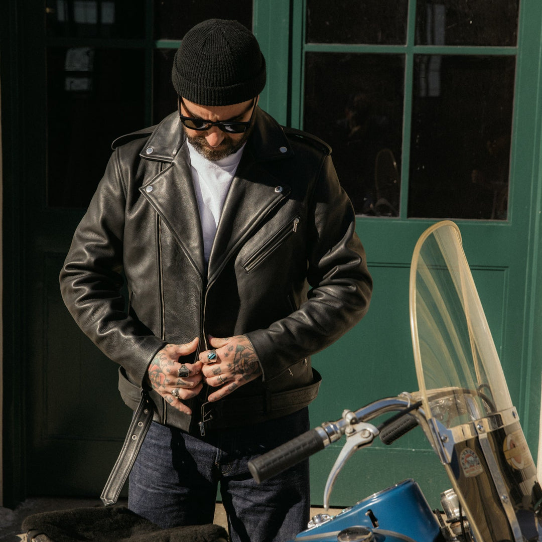 Man in leather jacket and sunglasses standing next to a blue motorcycle against a green door.