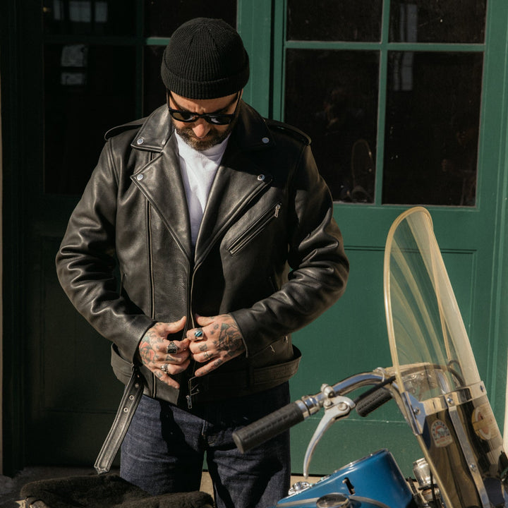 Man in leather jacket and sunglasses standing next to a blue motorcycle against a green door.