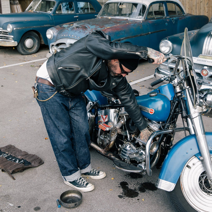 Person working on a blue motorcycle in a parking lot with vintage cars in the background